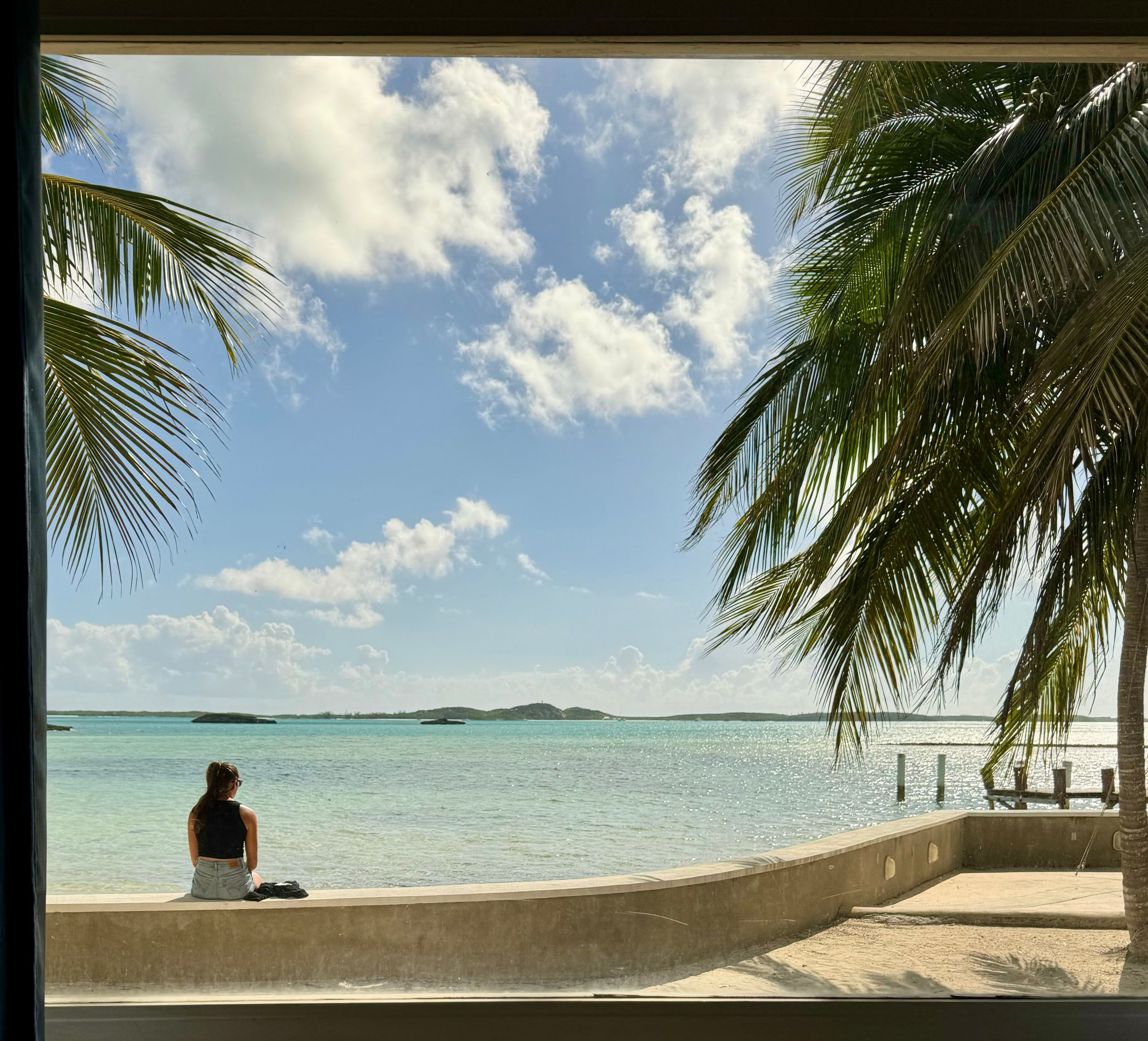 A person sits on the seawall looking across Elizabeth Harbour, with palm trees and the dock.