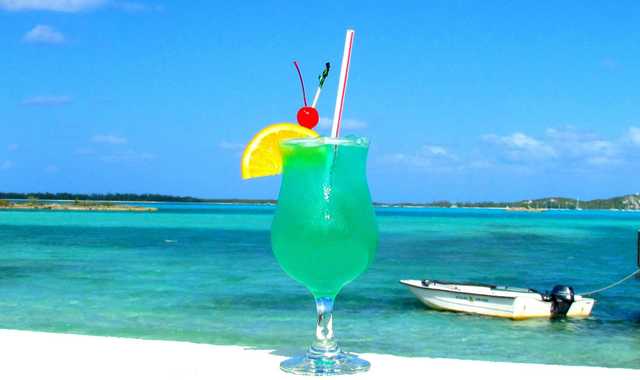 A turquoise drink with an orange slice and a cherry sits on a white ledge, with a small boat on the water behind it.